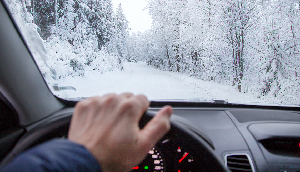 Крупним планом чоловік їде в лісі взимку по засніженій дорозі.Безпечне водіння на слизьких зимових дорогах вимагає зосередженості.Стаття AARP містить поради щодо водіння взимку.